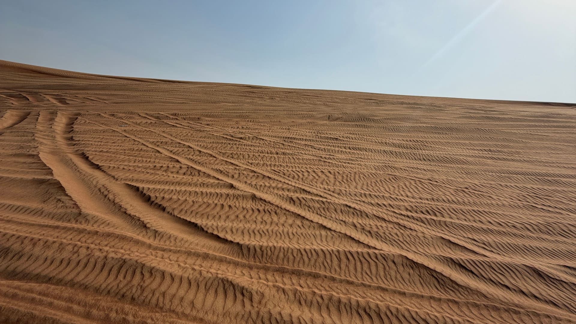Wind ripples and vehicle tracks across a sunlit dune slope under a pale blue sky