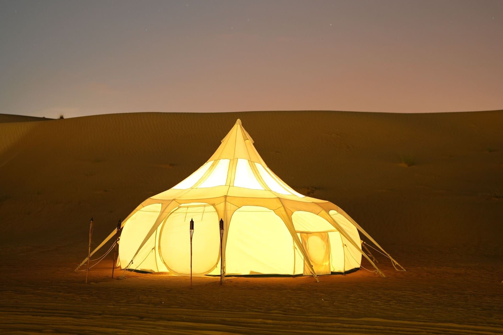 A glowing white tent with bamboo torches on a dark dune after sunset