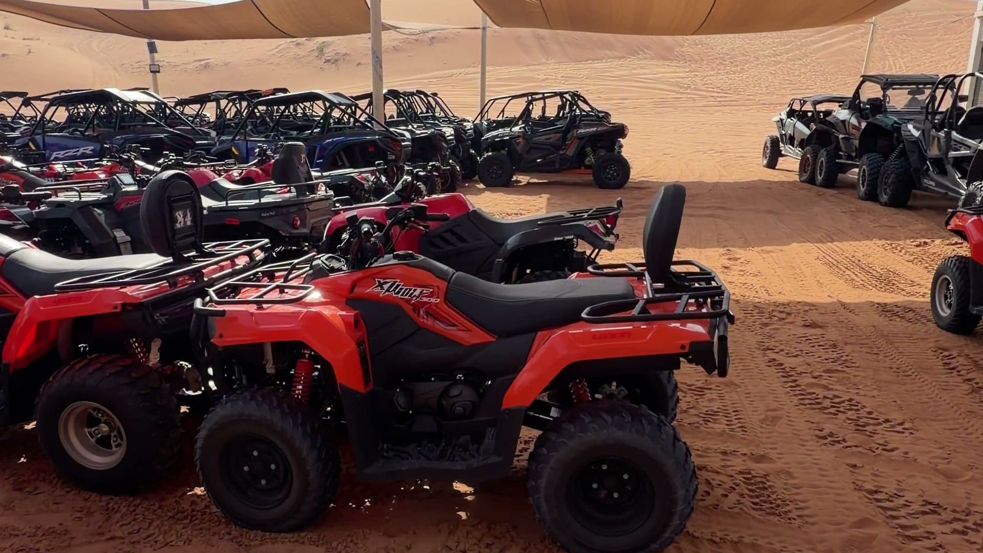 Rows of quad bikes and buggies parked under shade sails beside orange dunes